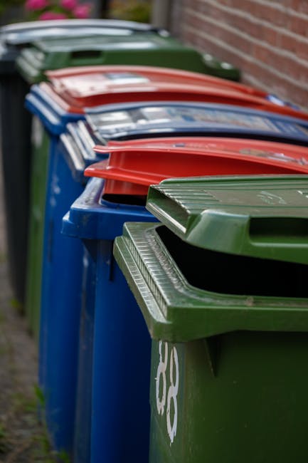 A row of multiple large wheelie bins lined up outdoors along a pavement beside a brick wall. The bins are predominantly blue with a green bin in the foreground and red lids on some of the blue bins. T